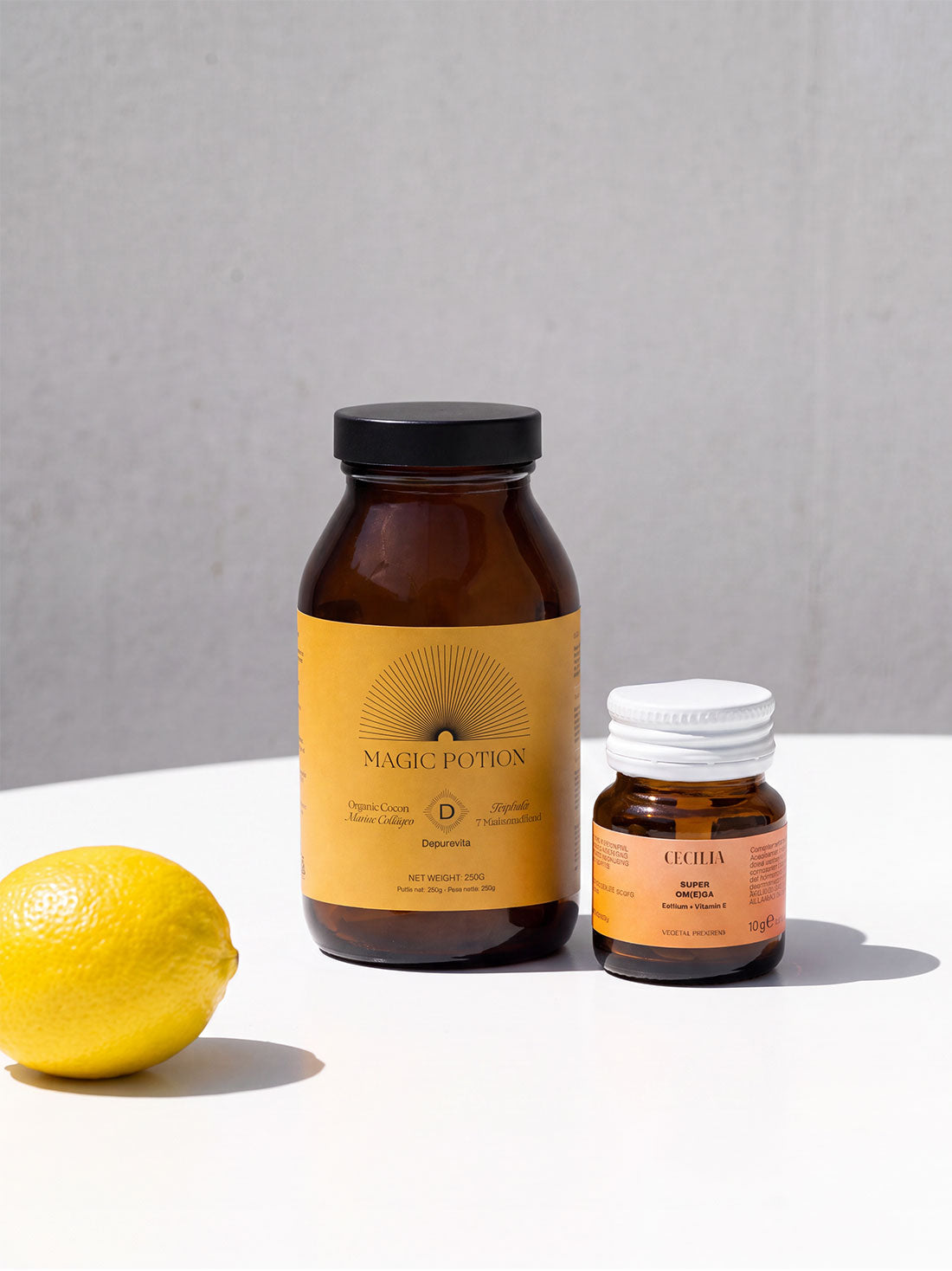 depuravita and super omega amber supplement bottles on a white surface against a light gray wall: a larger bottle labeled Magic Potion with a black lid and a yellow label, and a smaller bottle labeled ECHO with a white lid and orange label. A yellow lemon sits to the left.