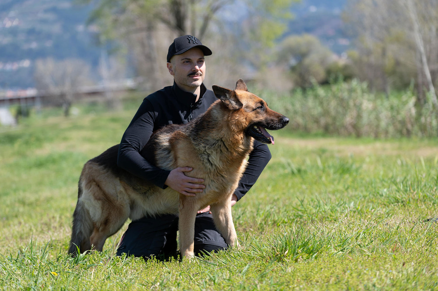 Andrea Fruzzetti reiki master in a black long-sleeve shirt and black cap kneels in a grassy field, holding a German Shepherd dog.