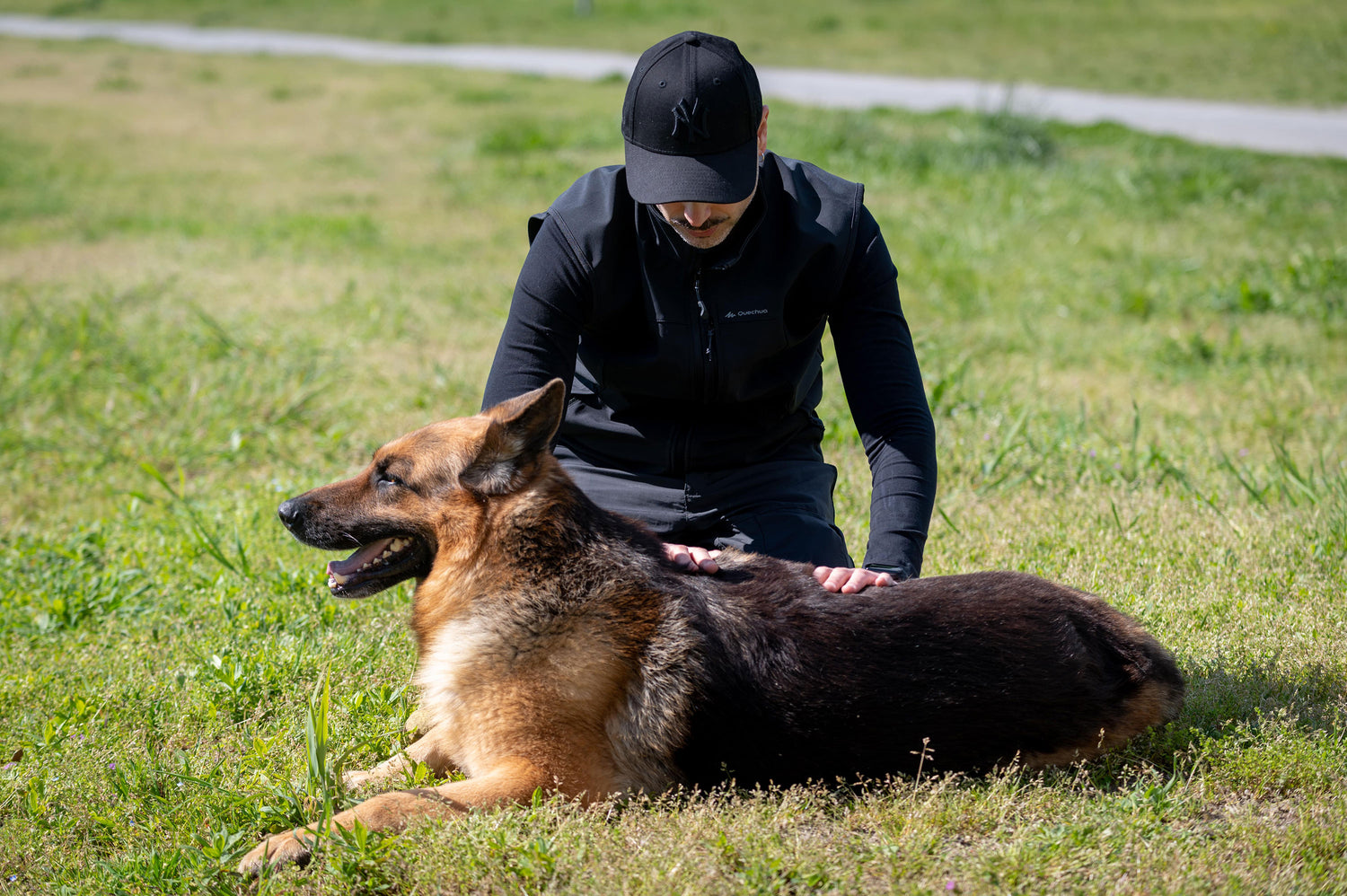 Andrea Fruzzetti Reiki master in a black cap and black long-sleeve top kneels on grass while petting a German Shepherd lying on the grass outdoors.