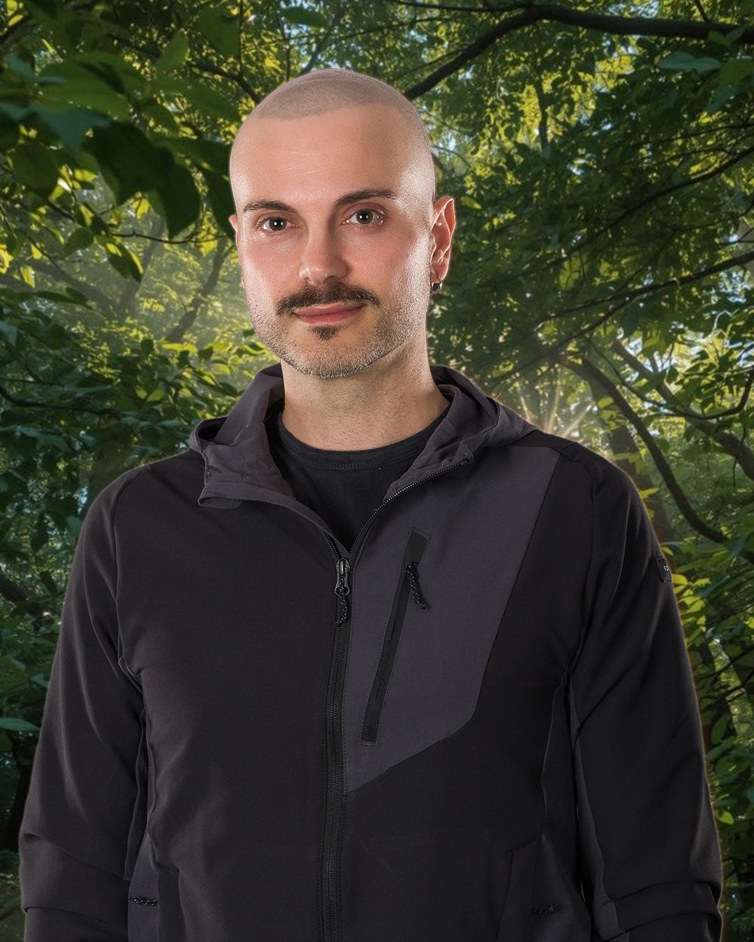Andrea Fruzzetti reiki master wearing a black and dark gray zip-up hooded jacket, standing outdoors in front of a background of green trees.