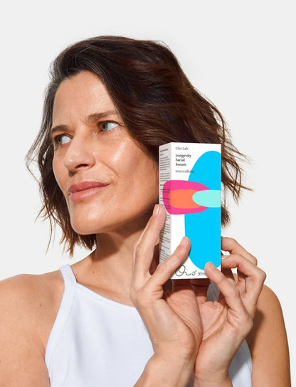 A woman with short brown hair holds a box of Oio Lab Intercellular Serum Longevity Fuel Intense Caffeine against a clean white background.
