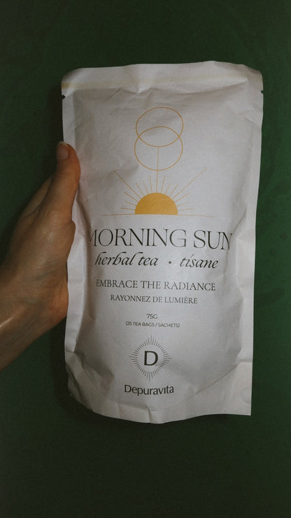 A hand holding a white stand-up pouch of Morning Sun herbal tea against a dark green background. The pouch features a minimalist gold sun and circle logo and the brand name Depuravita.