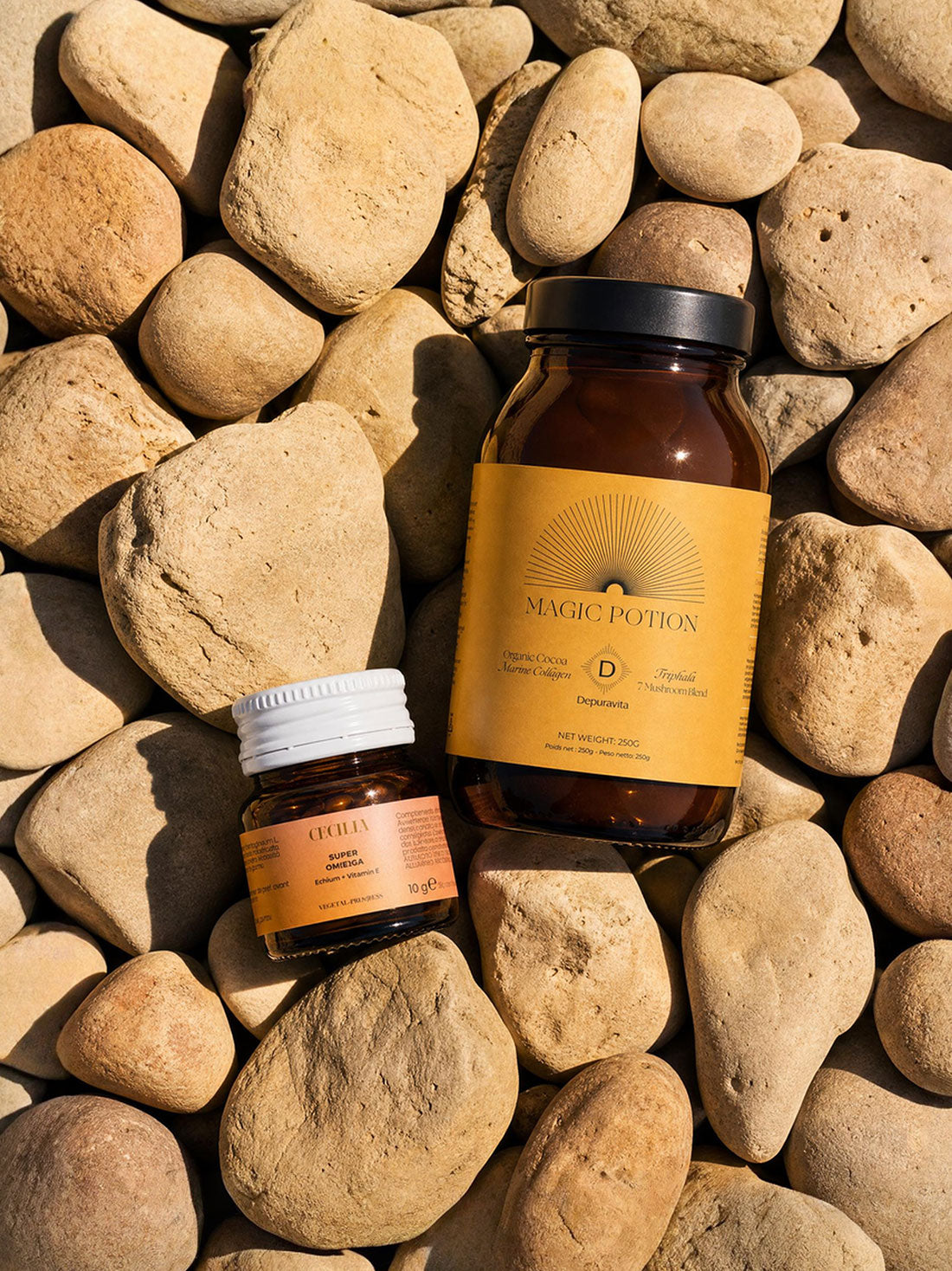 Two amber glass supplement bottles from Magic Potion, one larger bottle with a black lid and one smaller jar with a white lid, arranged on a bed of smooth tan river stones.