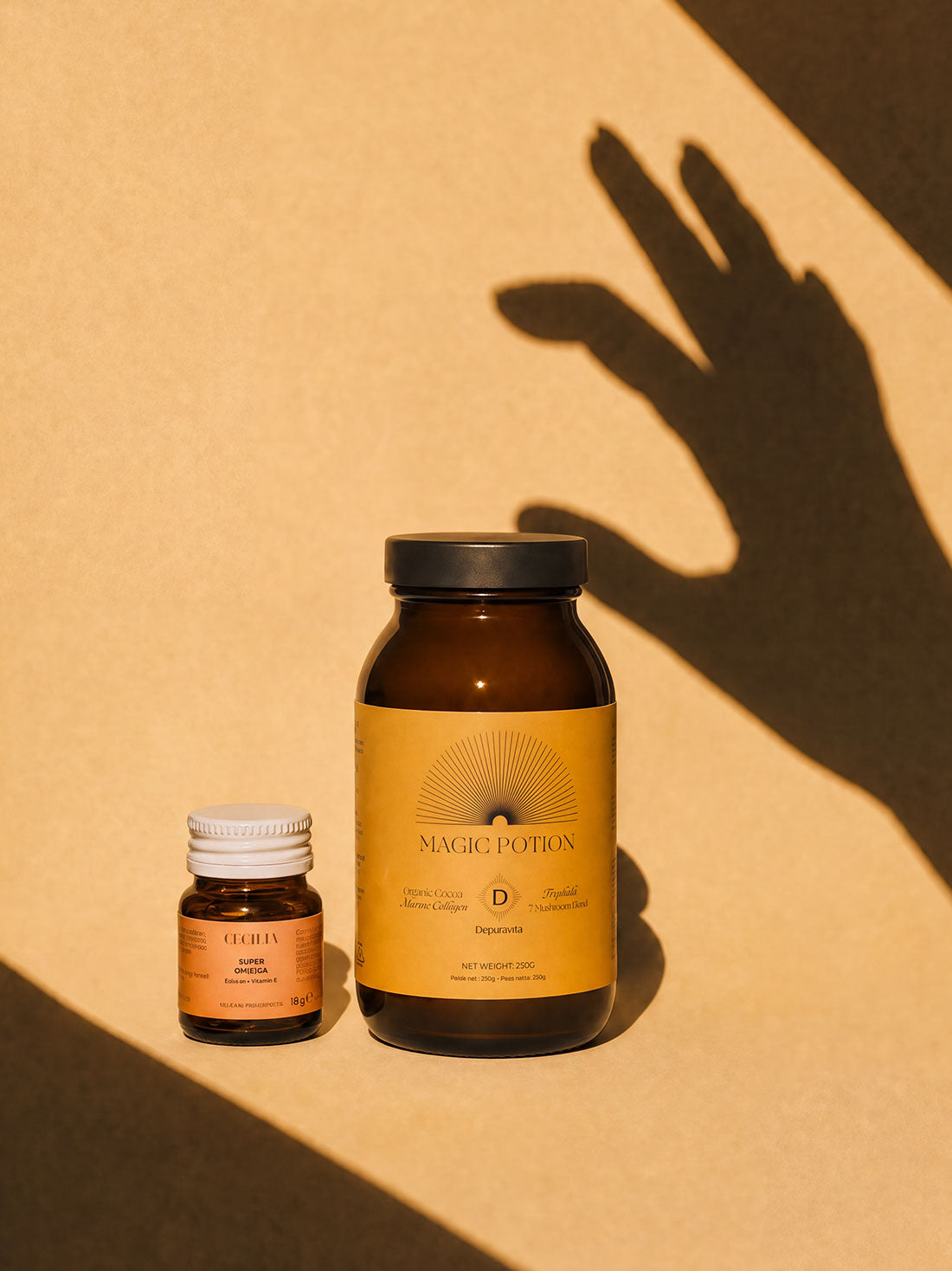 Two amber glass supplement bottles with yellow labels on a warm beige surface, cast in sharp shadow by a hand. The larger bottle reads Magic Potion and the smaller bottle reads Chakra.
