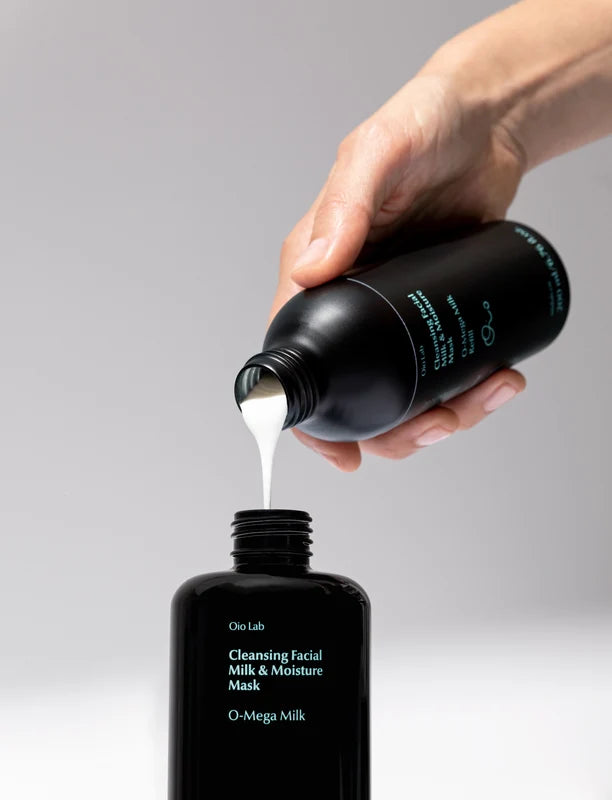 A hand pours white O-Mega Milk cleansing facial milk from a matte black bottle into another matte black bottle labeled &