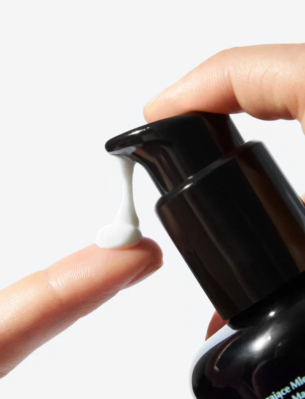 A close-up of Oio Lab O Mega Milk hand pressing a black pump dispenser, with a small dollop of white cream being dispensed onto another finger against a clean white background.