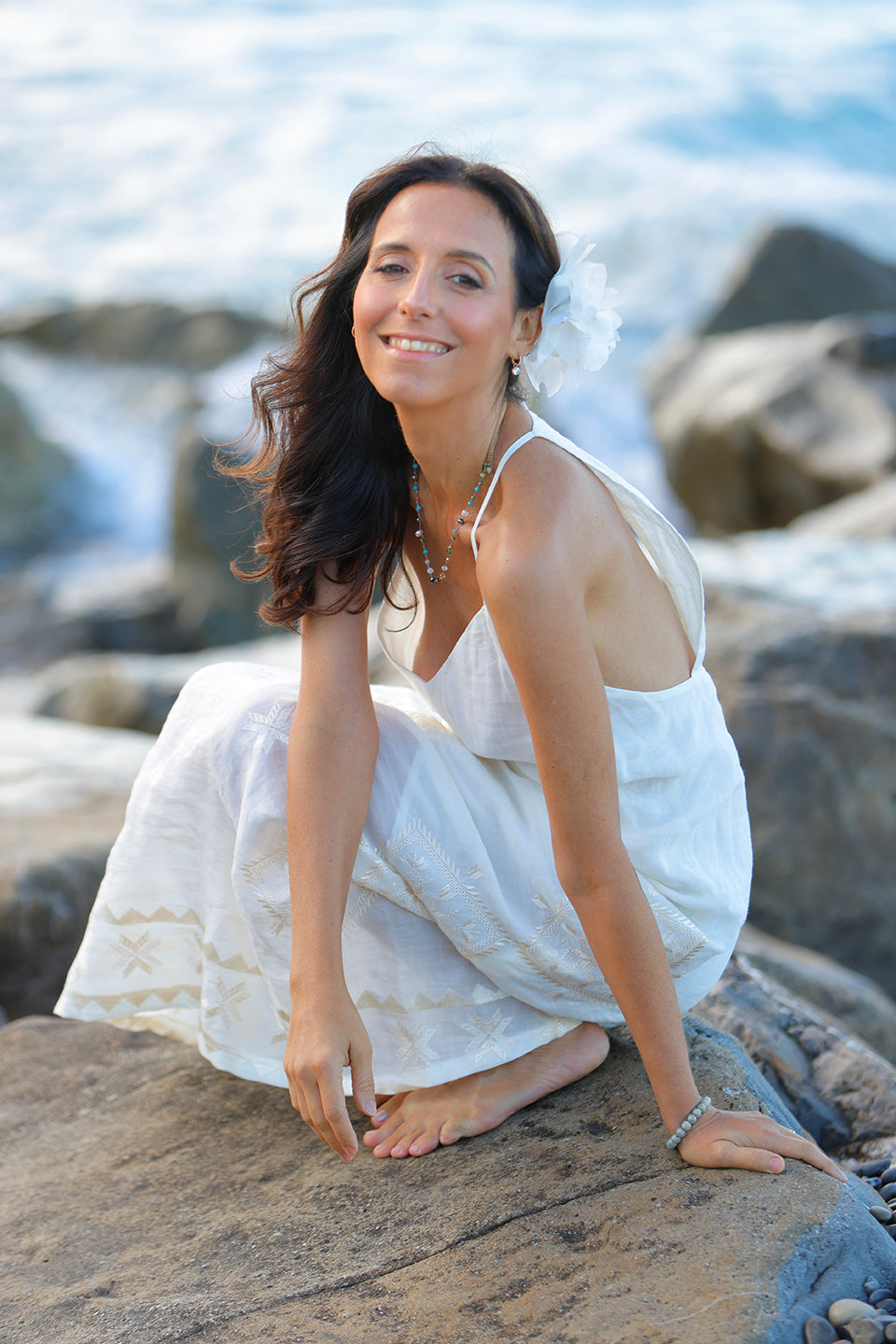 Benedetta Spada with long dark hair sits barefoot on a rocky beach, wearing a sleeveless white dress and a white flower in her hair, with ocean waves blurred in the background.