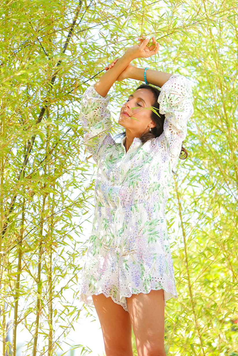 Benedetta Spada wearing a white crochet mini dress with a green and purple floral print, standing in a sunny bamboo grove with her arms raised.