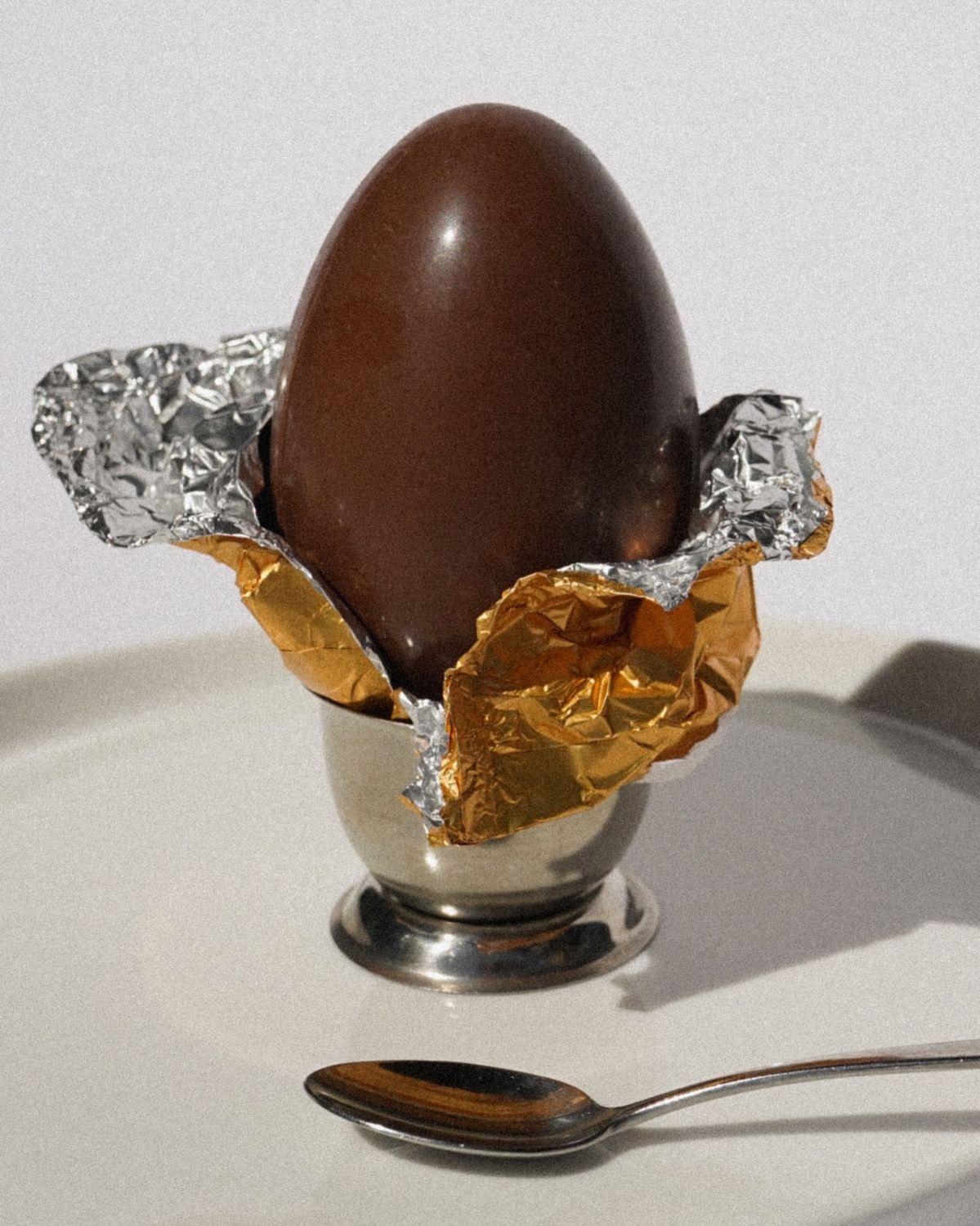 A glossy milk chocolate Easter egg resting in a silver egg cup surrounded by unwrapped gold and silver foil, representing the peak of the commercial sugar season that lands precisely when the body is biologically primed to reset.