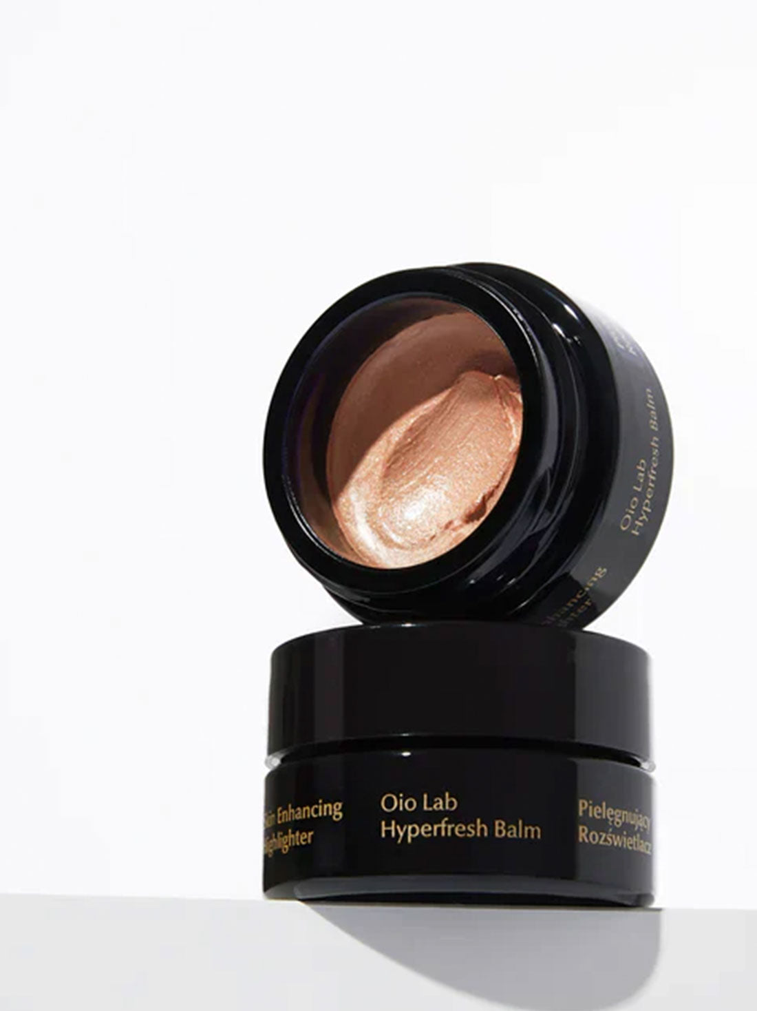 An open black cosmetic jar of Oio Lab Hyperfresh Balm with a metallic bronze highlighter balm visible inside, resting on top of a closed matching black jar against a clean white background.