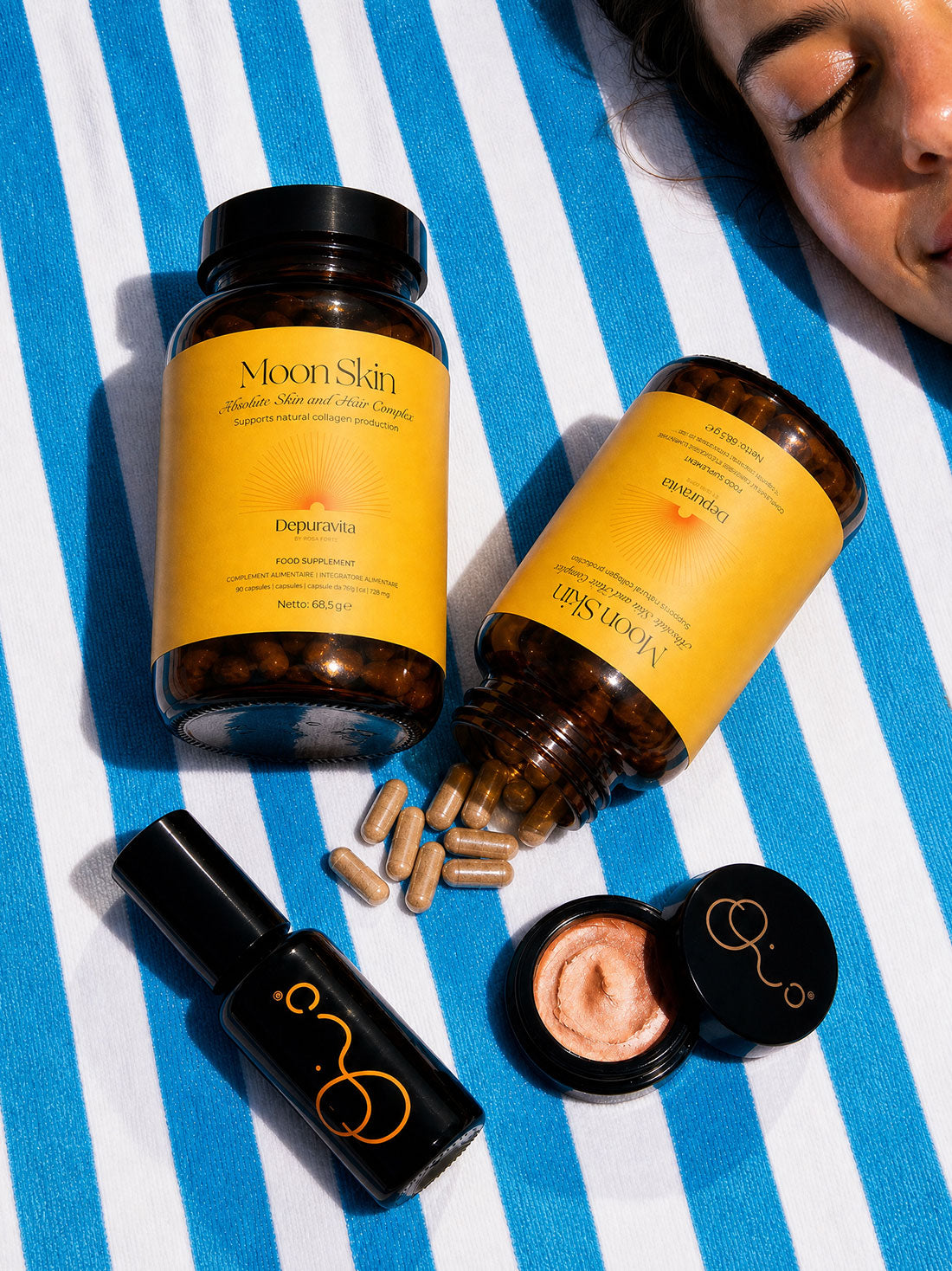 Two bottles of Moon Skin skincare products and a small black tube are arranged on a blue-and-white striped blanket, with one bottle tipped over to spill capsules. A woman&