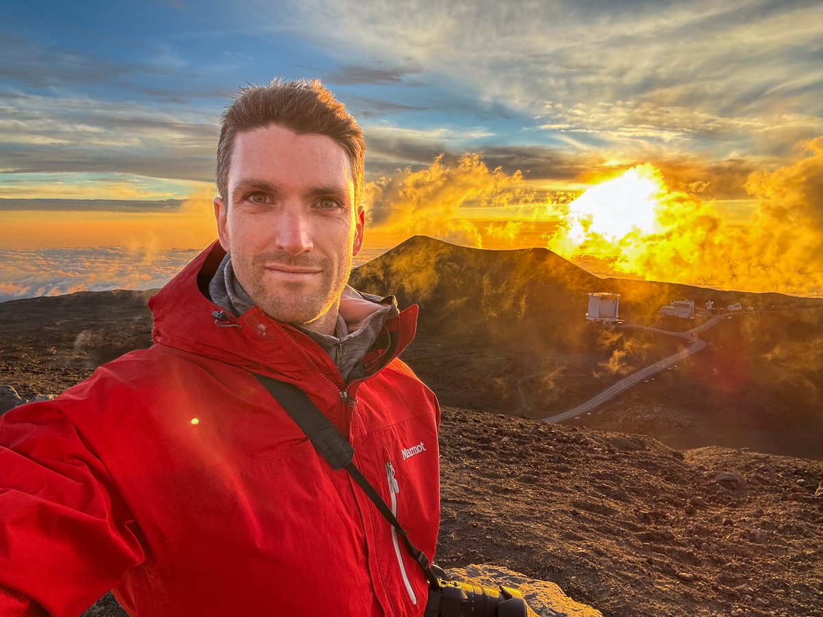 geophysicist stefan burns wearing a red hooded jacket takes a selfie on a mountain peak during a dramatic golden sunset, with clouds, steam, and a winding road in the background.
